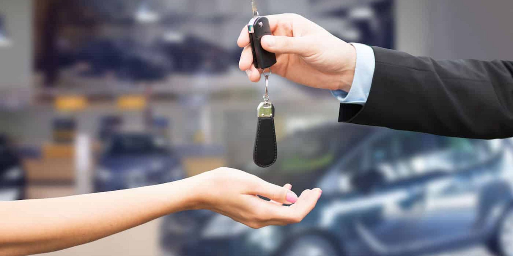 Eine Hand übergibt einen Autoschlüssel an eine andere Hand vor einem unscharfen Hintergrund mit Autos.